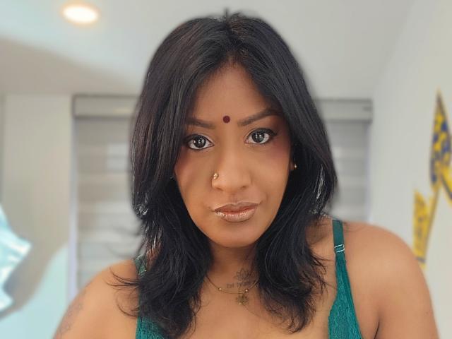 Nahirr with medium brown skin, black shoulder-length hair, nose ring, red dot on forehead, shiny bronze lipstick, green lace top, and gold necklace, looking directly at camera.