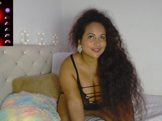 YiyaHot69 with long curly brown hair, tan skin, and black strappy lingerie, sitting on a bed, smiling, showing her boobs. A fluffy pillow and a colorful blanket are in front.