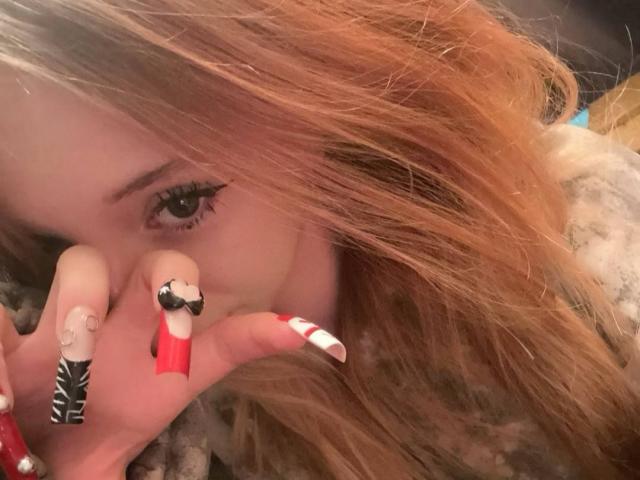 RinaChanofsky's face with red hair, black and white nail art, and red nail, showing her eyes and fingers, lying on a fuzzy gray blanket.