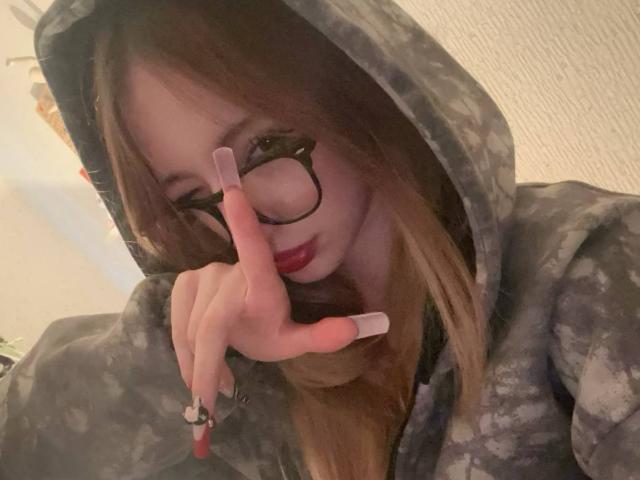 RinaChanofsky with long brown hair, glasses, and red lipstick, wearing a grey camo hoodie, covering her mouth with a hand showing long, white-painted nails.