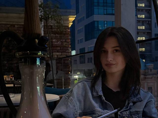 CharleenTilton with dark, shoulder-length hair, wearing a denim jacket, sitting near a glass hookah, smiling softly at the camera, in a dimly-lit urban setting.