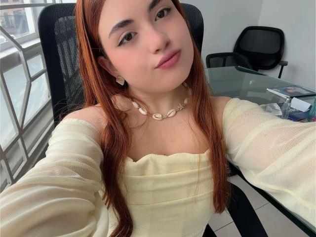 VicluX, con lunghi capelli rossi, pelle chiara e labbra rosa, indossa una maglietta gialla con una spalla scoperta e una collana di conchiglie, e sta scattando un selfie seduta su una sedia da ufficio..
