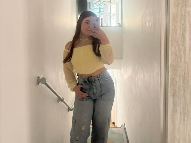 VicluX, con lunghi capelli castani, una maglietta gialla a spalle scoperte e jeans blu a vita alta e strappati, sta scattando un selfie allo specchio, mostrando un sedere carino, slanciato e sensuale, in un corridoio bianco..