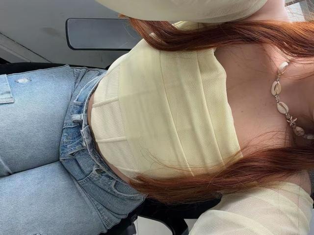 VicluX with red hair, yellow crop top showing tits, blue jeans, and a seashell necklace, taken from a low angle in a car. VicluX's ass is prominent.