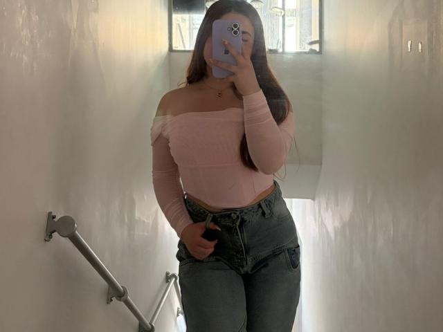 VicluX with long brown hair, pink off-shoulder top, and high-waisted blue jeans, taking a mirror selfie, showing her curvy ass and small tits, in a white hallway.