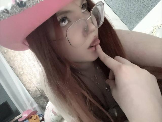 MargSeyler with long red hair, wearing glasses, pink hat, and finger in mouth, showing off her tits, pale skin, and a cute, sexy look.