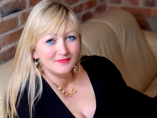MarinaSweet has blonde hair, blue eye makeup, pink lipstick, black top, gold necklace, and is smiling with her tits slightly visible, sitting on a beige leather couch.