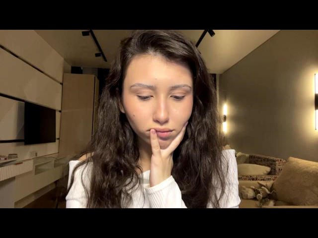 NecoleArciniega with long wavy brown hair, white shirt, and finger touching her lips, looking down. She has a small mole on her right cheek, soft brown eyes.