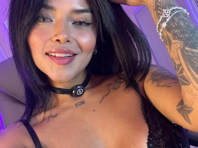 MajoJones has long black hair, tattoos on her arm and chest, and is wearing a black lace top with a collar. She's smiling with pink lipstick and a bracelet.