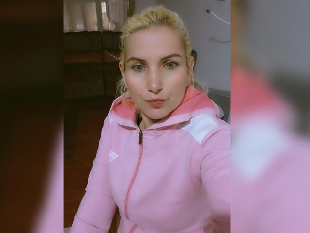 GiuliaAddams, con capelli biondi, occhi verdi e una felpa rosa con zip, con un'espressione leggermente accigliata, tiene in mano un selfie con uno sfondo rosa e bianco sfocato. Il suo seno è parzialmente visibile..