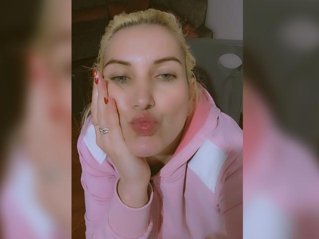 GiuliaAddams fronce les lèvres, ses lèvres roses brillantes, et porte un sweat à capuche rose avec des rayures blanches, une main sur la joue, les cheveux blonds attachés en queue de cheval, des ongles rouges et une bague au doigt..