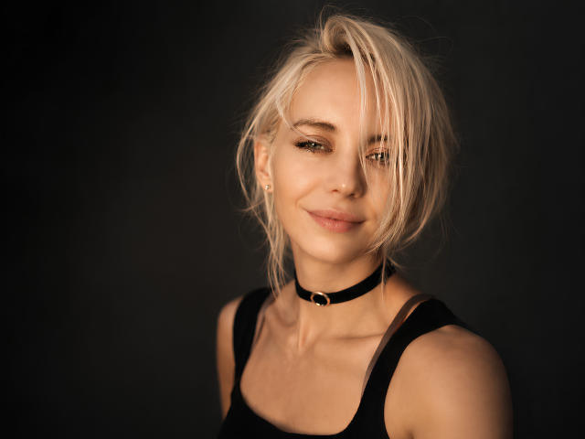 BlondIceX has blonde hair, a black choker, and a slight smile. She's wearing a black tank top, showing her tits, with a soft, sexy, and confident look.