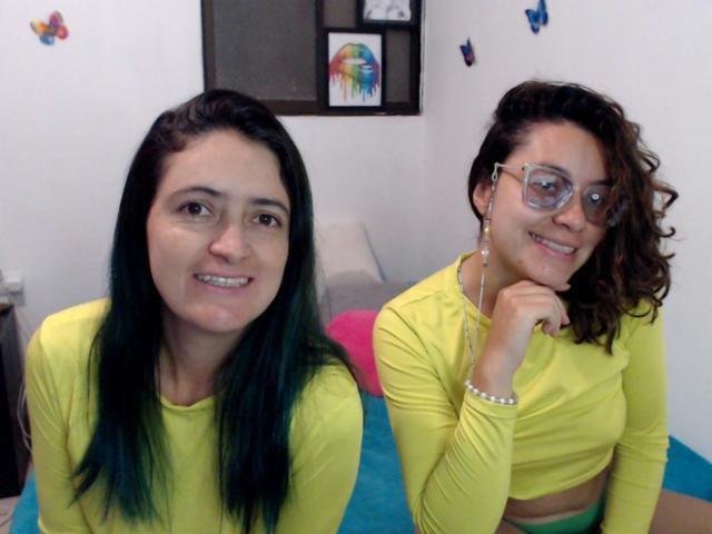 LunaVenus with green-tipped black hair and LunaVenus with curly brown hair and glasses, both wearing yellow tops, sitting on a blue bed, smiling. LunaVenus has small tits.