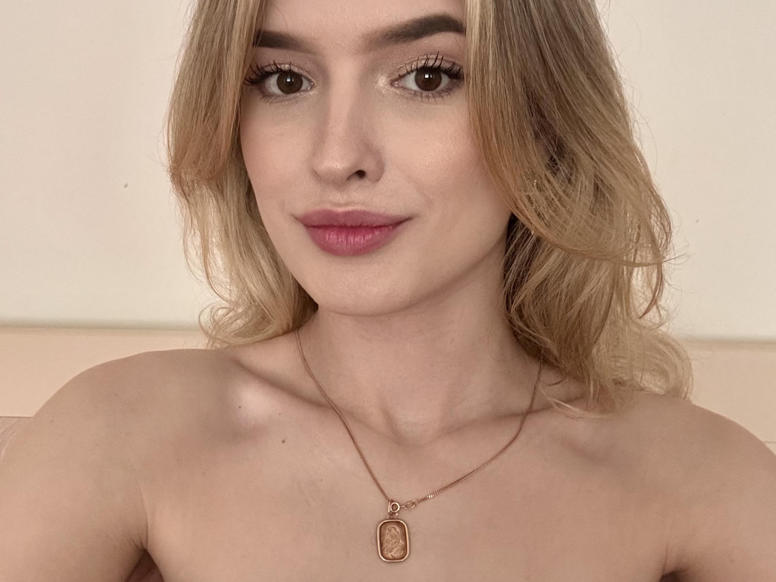 JoleenVigier, sem blusa, loira, cabelo ondulado, olhos castanhos, batom rosa, maquiagem discreta, nariz pequeno, colar com pingente em formato de placa, sorrindo suavemente, pele clara, fofa, atraente, foto da parte superior do corpo..