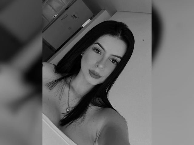 Black-and-white selfie of IsabellaJames with long dark hair, wearing a strapless top, showing her tits slightly, looking into the camera with a neutral expression, necklace visible.