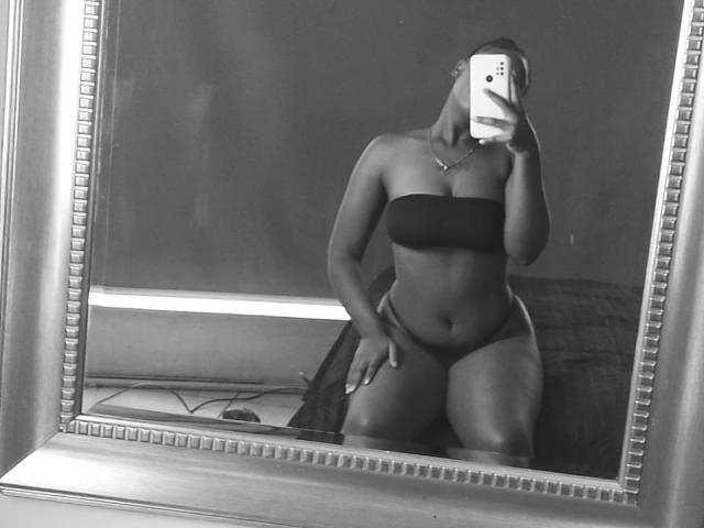 LilyBrownHot, con la pelle scura, indossa un top nero senza spalline e biancheria intima abbinata, seduta di fronte a uno specchio, tiene in mano un telefono, mostrando il sedere e la vulva..