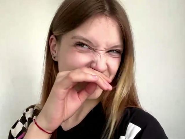 VeraBrabazon, with dirty blonde hair, is making a disgusted face, covering her nose with her hand, wearing a black and white shirt, and a red bracelet on her wrist.