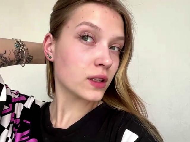 Vera Brabazon, mit heller Haut, grünen Augen und rosafarbenem Lippenstift, hat langes blondes Haar, einen tätowierten Arm und ein schwarz-weiß kariertes Hemd. Sie blickt mit leichtem Schmollmund in die Kamera..