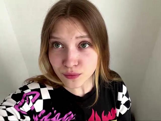 VeraBrabazon has green eyes, light skin, and straight brown hair. She's wearing a black and white checkered shirt with pink and purple designs. She looks surprised.