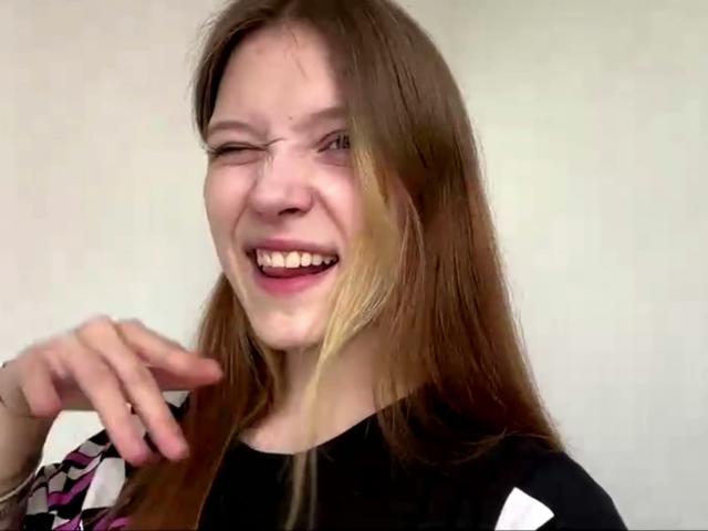 Vera Brabazon tem cabelos castanhos longos, pisca e sorri mostrando os dentes. Ela está vestindo uma blusa preta com mangas estampadas em branco e rosa. Sua mão direita está perto do rosto..