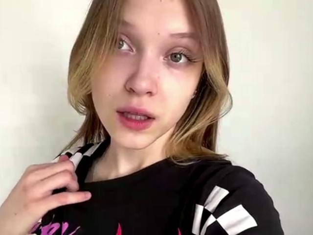 VeraBrabazon with light brown hair, green eyes, and pink lips, wearing a black and white checkered shirt, touching her collarbone with a slightly open mouth.
