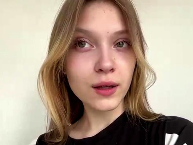 Vera Brabazon hat eine helle Haut, grüne Augen und blondes Haar mit rosafarbenen Lippen. Sie trägt ein schwarzes Hemd mit weißen Streifen und blickt direkt in die Kamera..
