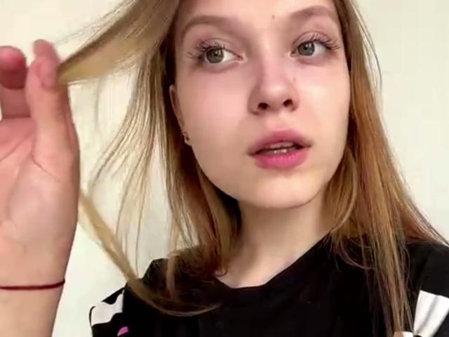 Vera Brabazon, avec des cheveux bruns clairs, tenant une mèche, des lèvres roses, des yeux verts, une chemise noire, un bracelet rouge, regardant légèrement sur le côté, une peau claire, un maquillage discret et une expression neutre..