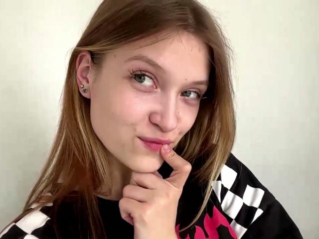 VeraBrabazon with light skin, green eyes, and brown hair, touching her pink lips with her finger, wearing a black and white checkered shirt, has a small nose piercing.