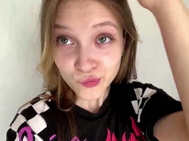 Vera Brabazon ha gli occhi verdi, il rossetto rosa e una camicia a quadri nera con scritte rosa. Sta facendo la smorfia e alzando la mano destra vicino alla testa..