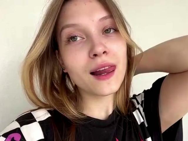 VeraBrabazon has blonde hair, green eyes, and is sticking her tongue out with pink lipstick. She's wearing a black-and-white checkered shirt, right arm raised.