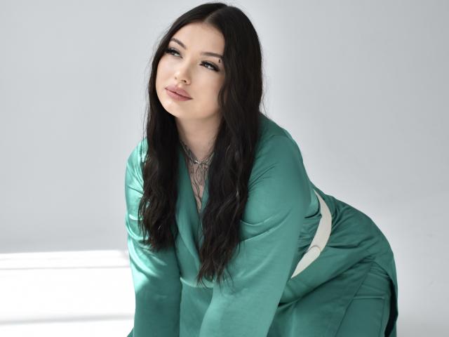 AsolMung with long dark hair, wearing a green dress, leaning forward, showing her tits slightly, with a confident, sultry expression, and a silver necklace.