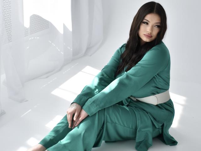 AsolMung has long dark brown hair, green scrubs with a white belt, and is sitting with a slight smile, looking at the camera, showing her ass.