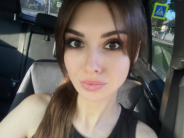 KylieJoys heeft volle, roze lippen, bruine ogen met dikke wimpers, en bruin haar in een staart, ze draagt een zwarte top en zit in een auto met een veiligheidsgordel..