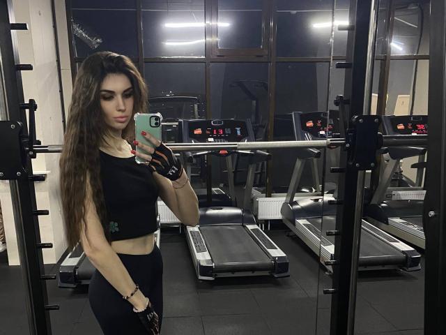 KylieJoys, con lunghi capelli castani, reggiseno sportivo nero e leggings neri, che tiene un telefono in un selfie allo specchio in palestra, mostrando il sedere e indossando guanti senza dita..