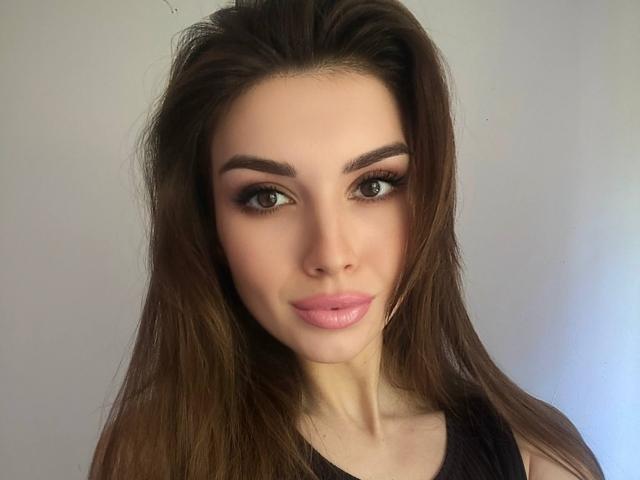KylieJoys tiene el pelo largo y castaño, cejas pobladas, lápiz labial rosa y ojos marrones. Lleva una blusa negra, y tiene un rostro ligeramente sonrojado y adorable..