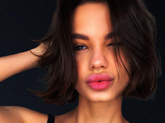 MiraPics-hot heeft golvend bruin haar, één oog gesloten en roze lippenstift. Ze knipoogt met haar rechteroog, haar linkerhand raakt haar hoofd, tegen een donkere achtergrond..