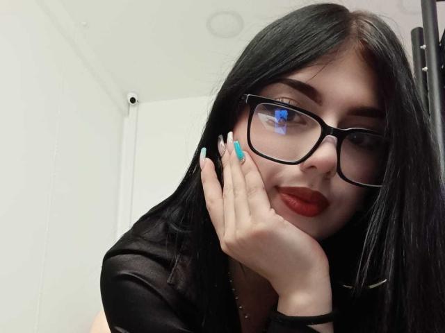 Isabella Rossi, con cabello negro, gafas, lápiz labial rojo y uñas azules, tocándose la mejilla con la mano, vistiendo una camisa negra, mirando a la cámara..