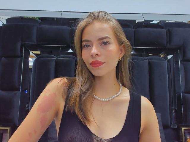 AnnaFer, avec du rouge à lèvres, portant un haut noir sans manches et un collier de perles, se tient devant des sièges de bus noirs, montrant une ecchymose rouge sur le haut de son bras droit..