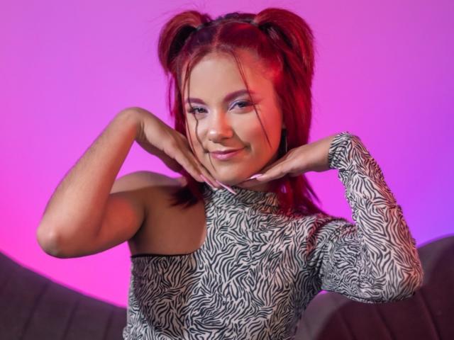 SkylerRouse with red pigtails, wearing a zebra-patterned dress, hands on cheeks, smiling, pink and purple lighting, showing off cute tits and a confident, sexy look.