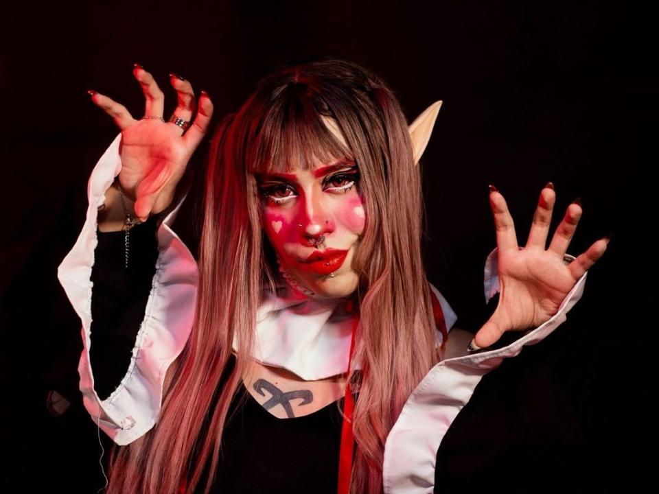 BonnyYoshida with long pinkish-brown hair, elf ears, red heart makeup, black outfit, white collar, raised hands, red nails, dark background, looking intense.