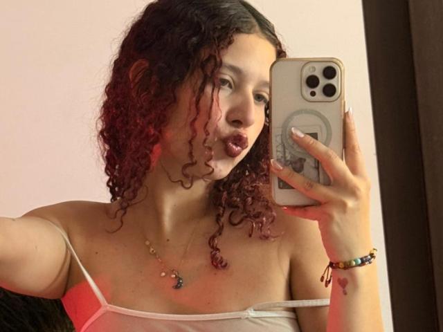 ValeryScot with curly red-tinted hair, pouty lips, wearing a white spaghetti strap top, holding a phone, necklace, and bracelet, taking a mirror selfie. Boobs visible.