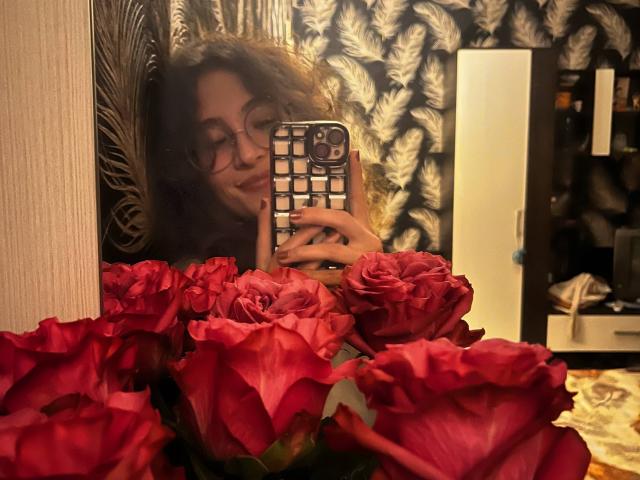 AlyceErway mit lockigem, braunem Haar und Brille, die ein Handy mit einer durchbrochenen Hülle hält, spiegelt sich in einem Spiegel wider, im Vordergrund befinden sich rote Rosen..