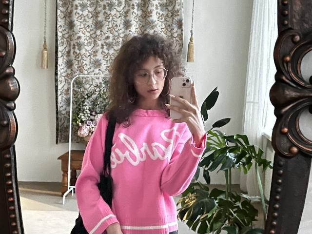 AlyceErway with curly hair, glasses, pink sweater with white "Adidas" logo, holding phone in mirror selfie, pink wristband, black backpack, standing in room with green plant.