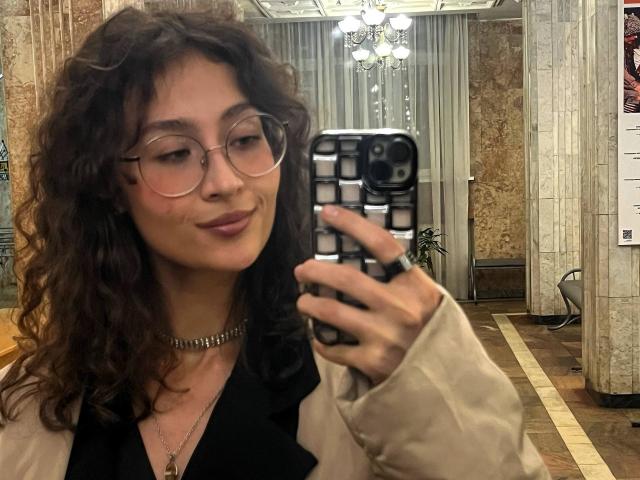 AlyceErway, avec ses cheveux bruns bouclés, ses lunettes, son collier et sa veste beige, prend un selfie dans un miroir, tenant un téléphone avec une coque noire à motifs, et souriant légèrement..