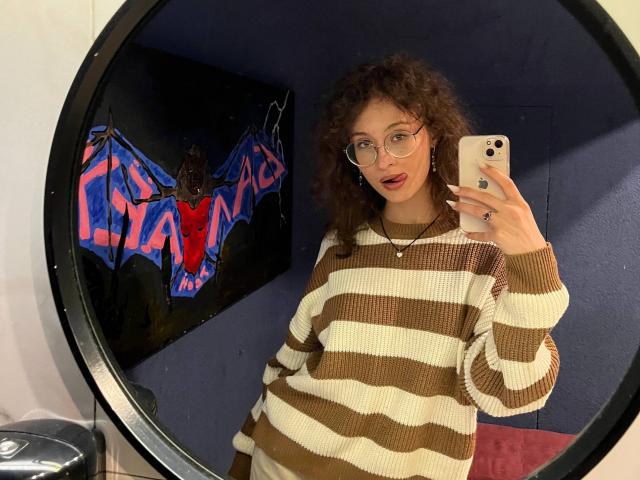 AlyceErway mit lockigem, braunem Haar, Brille und rotem Lippenstift, trägt einen braun-weißen gestreiften Pullover und macht ein Selfie mit ihrem Smartphone, wobei ihre Genitalien im Spiegelbild zu sehen sind..