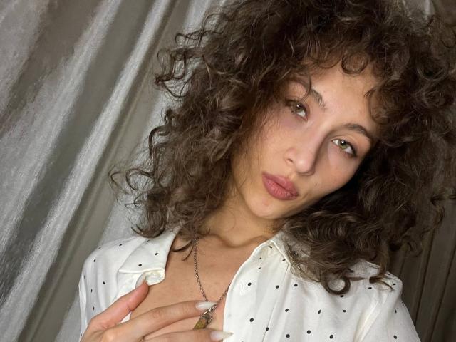 AlyceErway, avec ses cheveux bruns bouclés, ses lèvres roses et pulpeuses, et une chemise blanche à pois, tenant sa poitrine, portant un collier et regardant légèrement sur le côté..