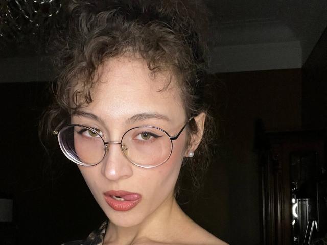 AlyceErway with curly brown hair, round glasses, light skin, and red lipstick, wearing a pearl earring, looking at the camera with a slightly open mouth.