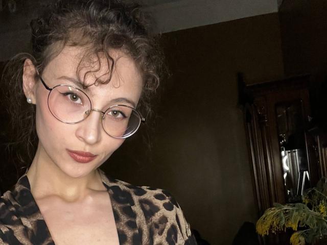 Alyce Erway, con cabello castaño rizado, usando gafas redondas y una blusa de estampado de leopardo, tiene los senos ligeramente visibles, un rostro lindo y una sonrisa sutil, en una habitación con poca luz..