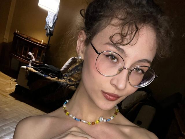 Alyce Erway, mit lockigem, braunem Haar, trägt runde Brillen und eine farbenfrohe Perlenkette. Sie hat ihre Hände auf ihrer nackten Brust, wobei ihre kleinen Brüste und der pinke Lippenstift sichtbar sind..