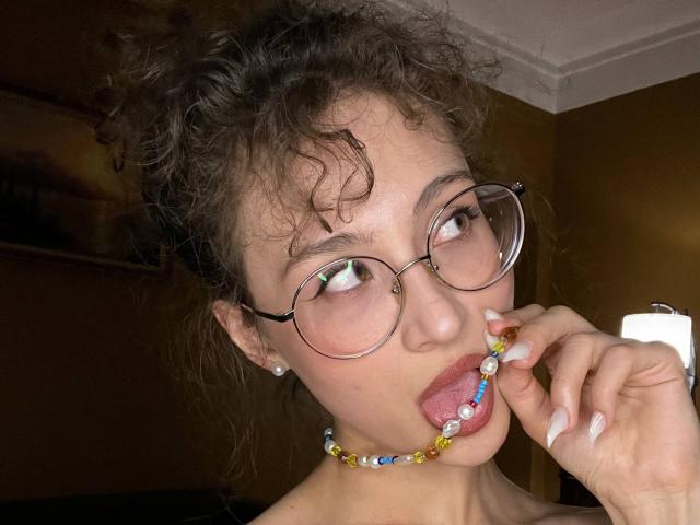 AlyceErway, com cabelos castanhos cacheados, óculos e um colar de contas, chupando contas coloridas, com a língua para fora, em um quarto com pouca luz, mostrando um piercing no nariz e brincos..